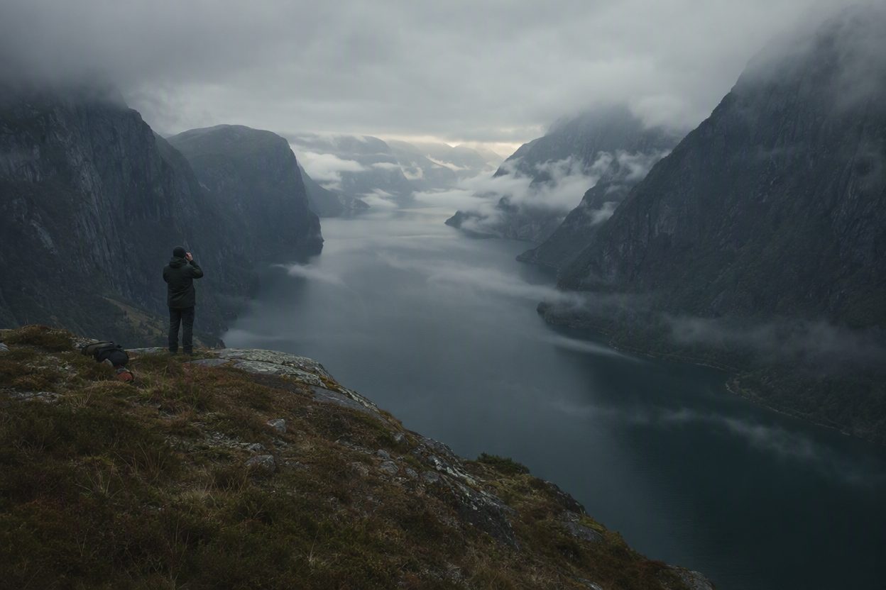 A lone figure on a high headland with binoculars, looking down the whole misty length of the fjord.